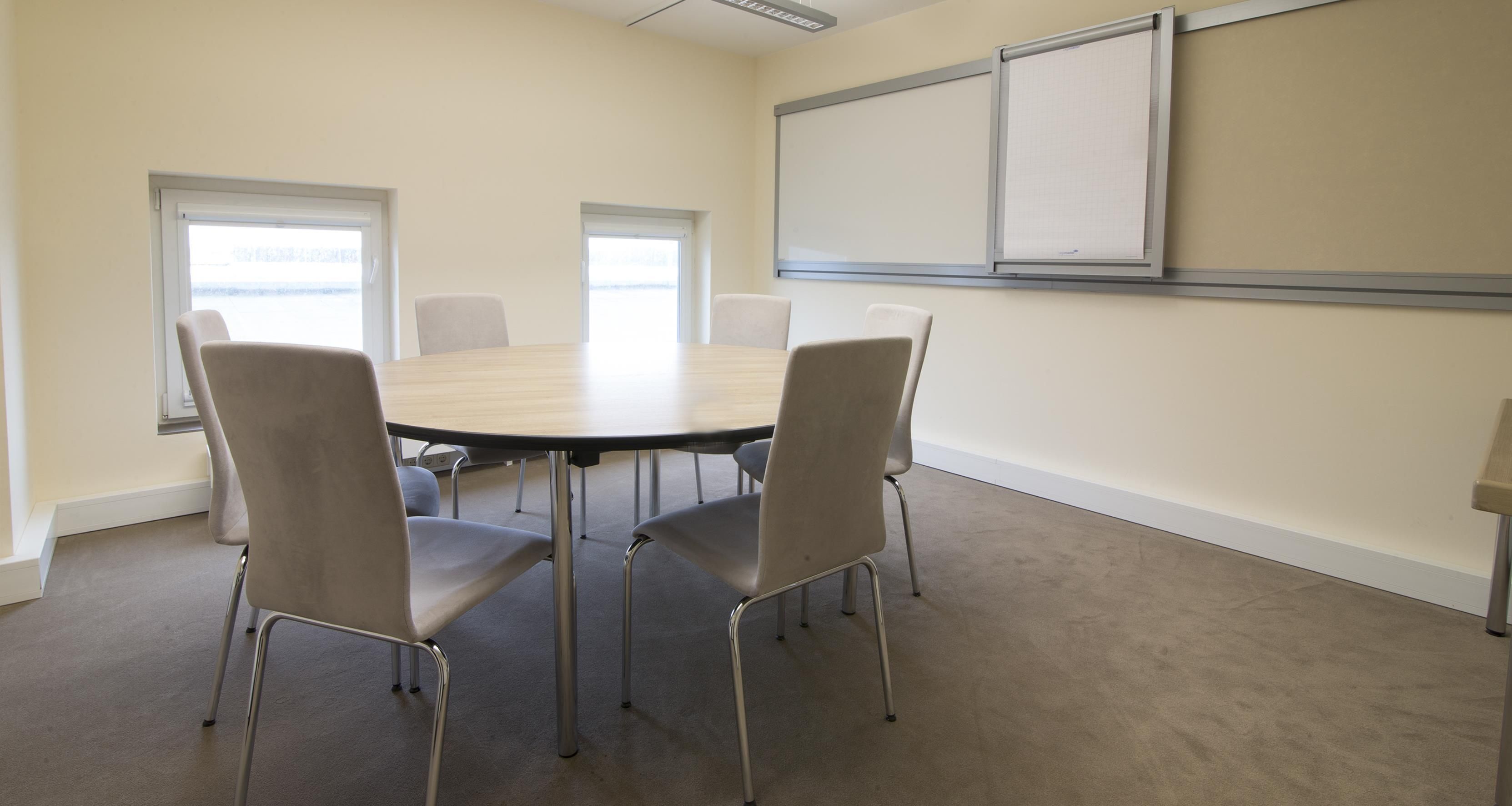 A conference room with a round table, four chairs, two windows, and a whiteboard on the wall.