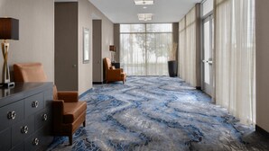 Eine Hotellobby mit einem fußbodenhohen Marmor-Muster, Leder-Sesseln, einer dunkelholzigen Kommode und großen Fenstern mit durchsichtigen Vorhängen.
