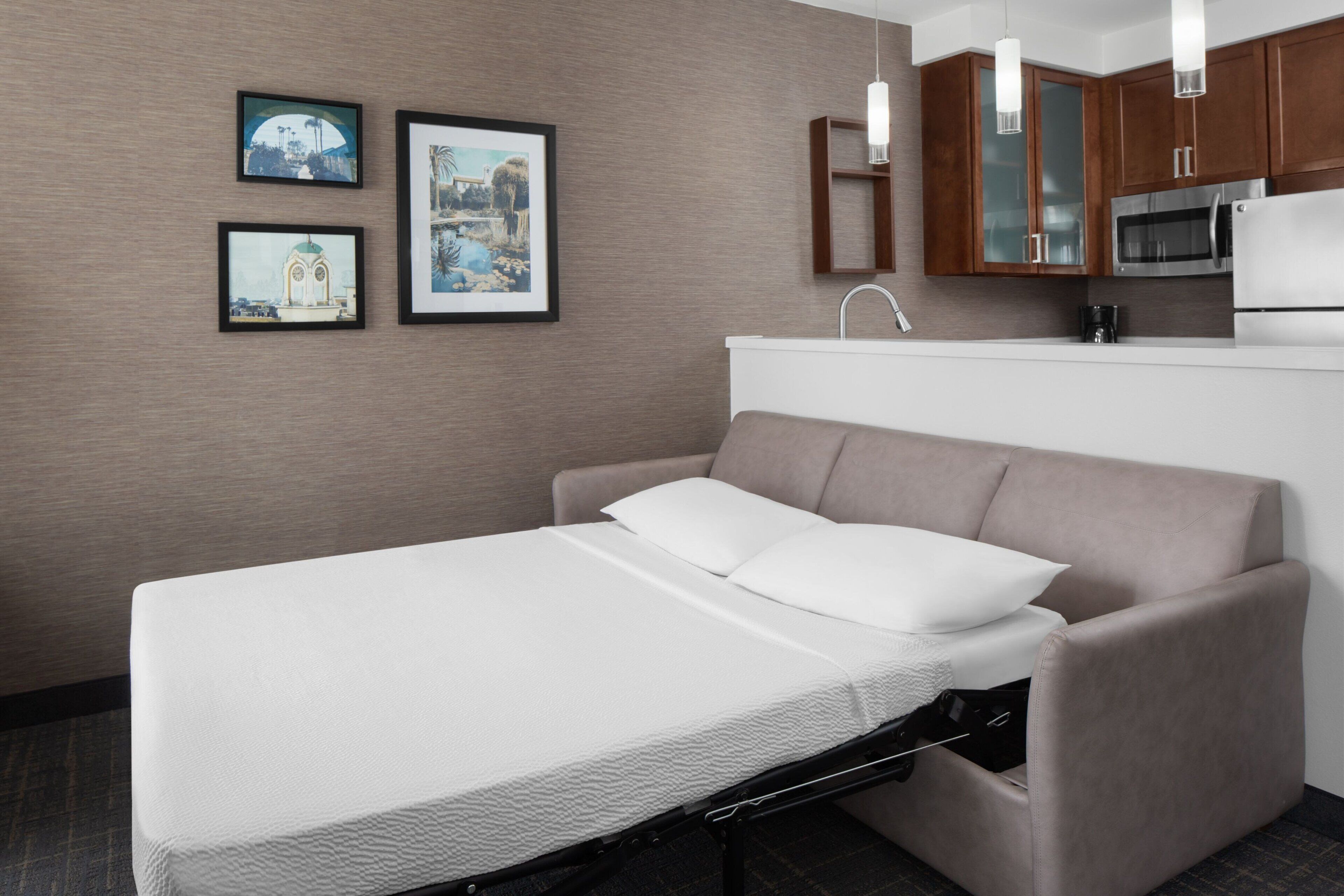 A compact hotel room with a bed, a kitchenette, and framed pictures on the wall.