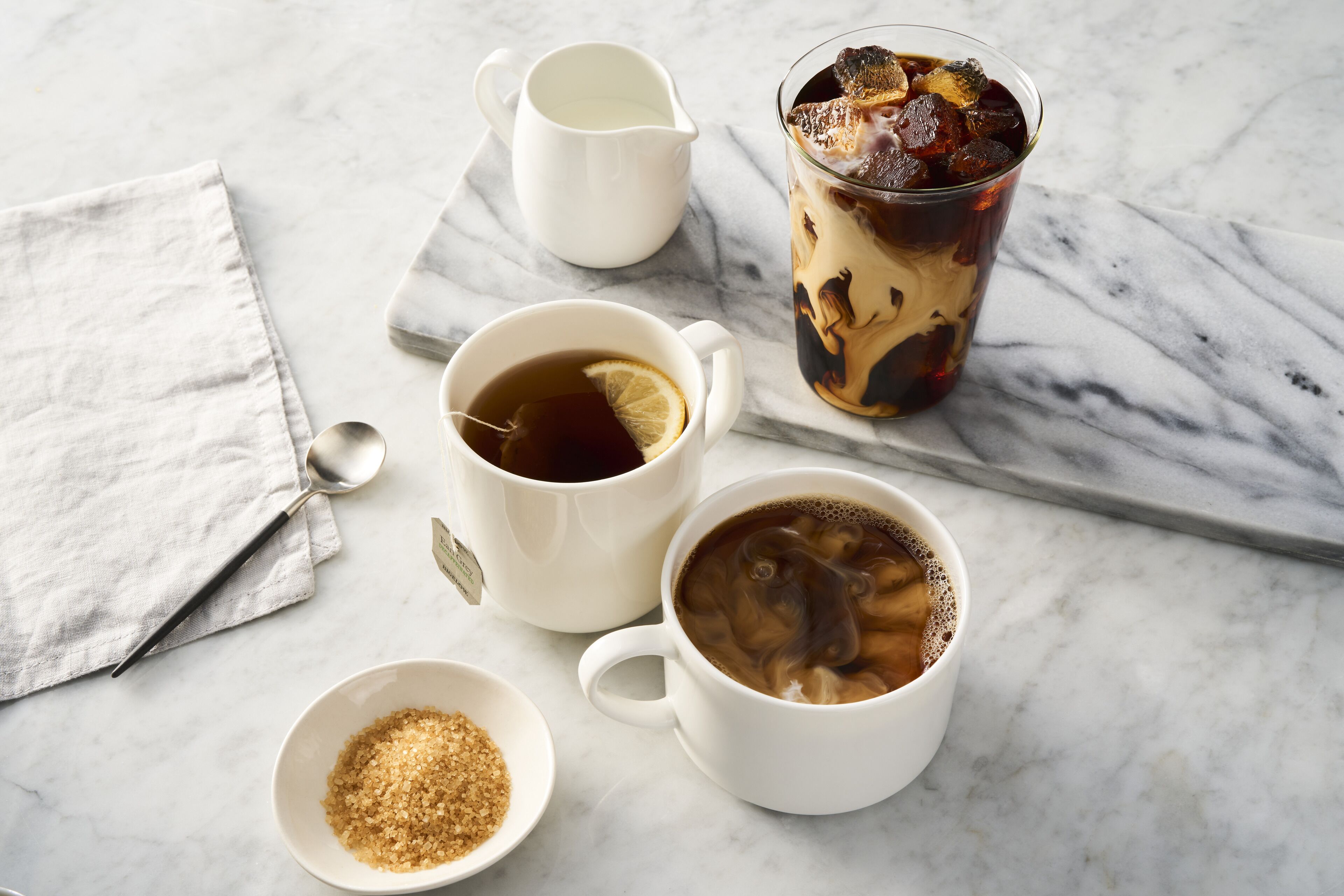 A marble table with two white cups of tea, a glass of iced tea, a small bowl of sugar, and a white pitcher.