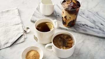 A marble table with two white cups of tea, a glass of iced tea, a small bowl of sugar, and a white pitcher.