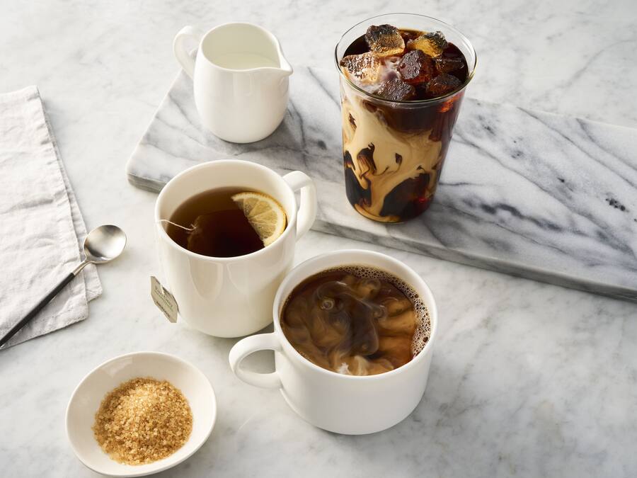 A marble table with two white cups of tea, a glass of iced tea, a small bowl of sugar, and a white pitcher.