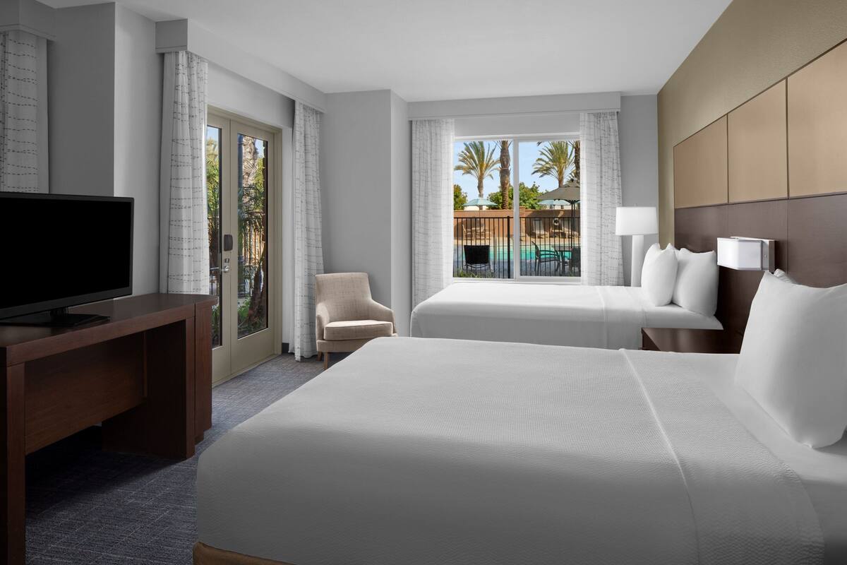 A hotel room with a large bed, a desk with a TV, a chair, and a window with a view of palm trees and a pool.