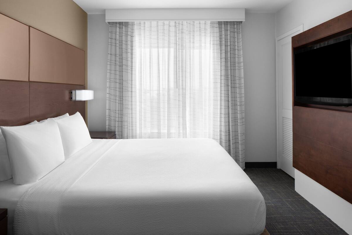 A modern hotel room with a large bed, a TV mounted on the wall, and a window with sheer curtains.