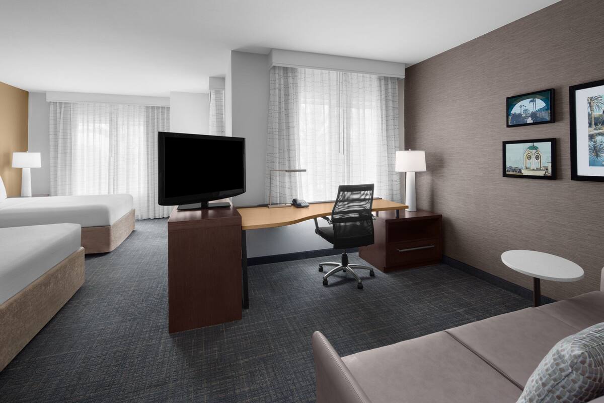 A hotel room with a bed, a desk with a computer, a television, a chair, a small table, and framed pictures on the wall.