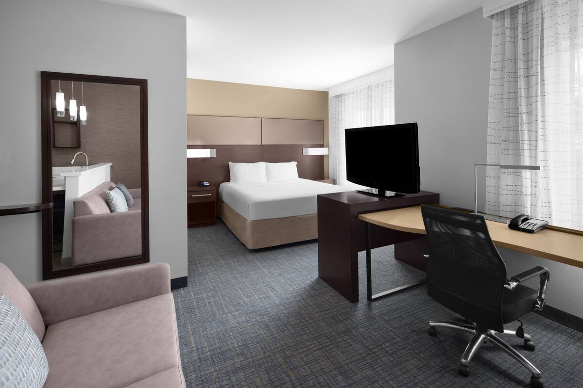 A hotel room with a bed, a desk, a chair, a TV, a mirror, a window with curtains, and a bathroom with a sink and mirror.