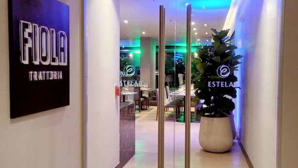 A hotel entrance with a glass door, a potted plant, and a sign reading "FIOLA TRATTORIA". - Hotel Estelar Calle 100 (Bogotá)