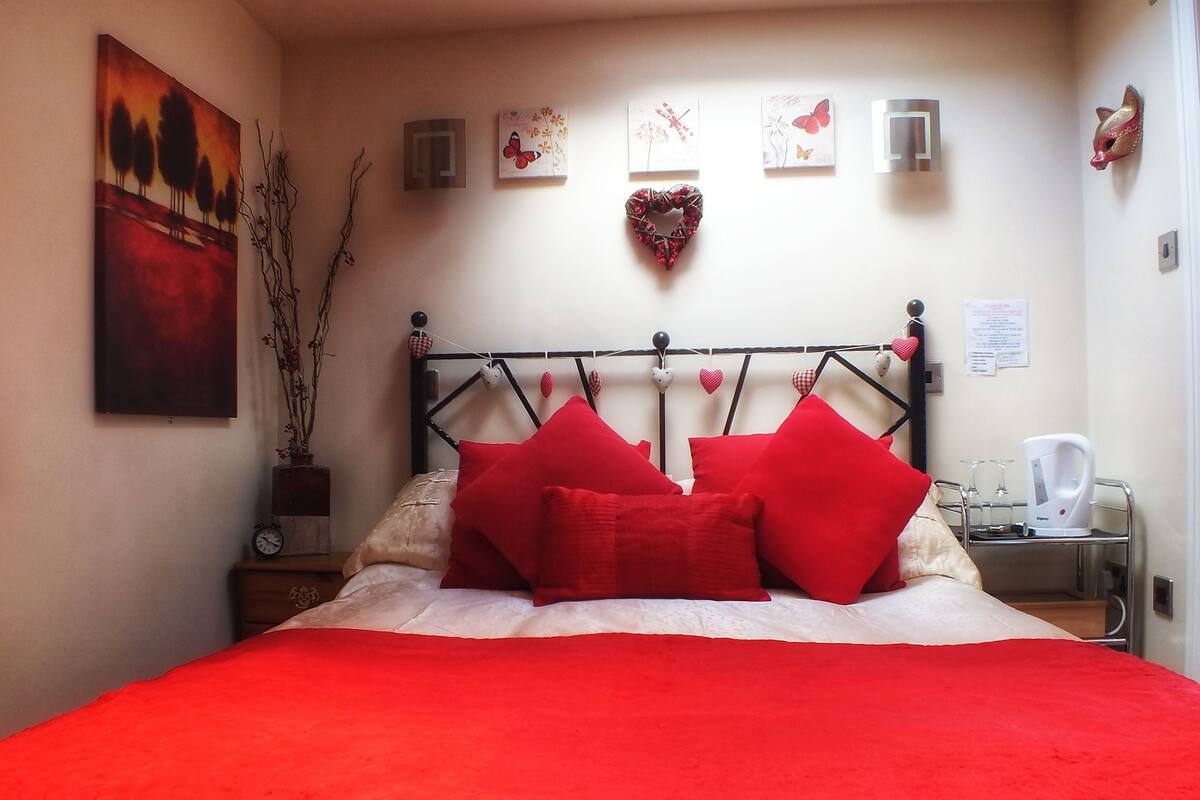 A bed with red bedding and pillows, a wall with artwork, a clock, and a kettle on a side table.