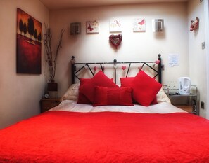 Ein Bett mit roter Bettwäsche und Kissen, eine Wand mit Kunstwerken, eine Uhr und ein Wasserkocher auf einem Nachttisch.