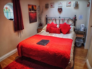 Un dormitorio con cama, ropa de cama roja, una mesita de noche, un reloj de pared y una ventana con cortinas.
