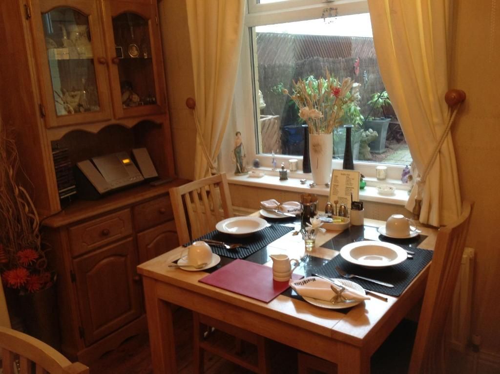 A dining area with a wooden table set for two, a window with curtains, and a view of the outside.