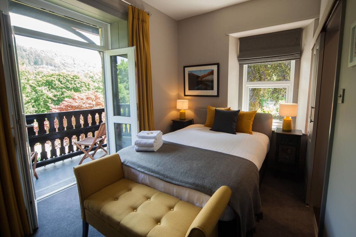 Une chambre d’hôtel comprenant un lit, un banc, une table de chevet avec une lampe, une fenêtre donnant sur des arbres et une porte menant à un balcon.