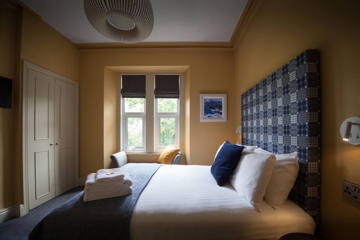 Une chambre d’hôtel avec un grand lit, une fenêtre avec des stores, un téléviseur fixé au mur et une tête de lit à motifs.