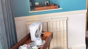 Habitación de hotel con televisor montado, bandeja de madera con cafetera y tazas, radiador y estante con un objeto con forma de corona.