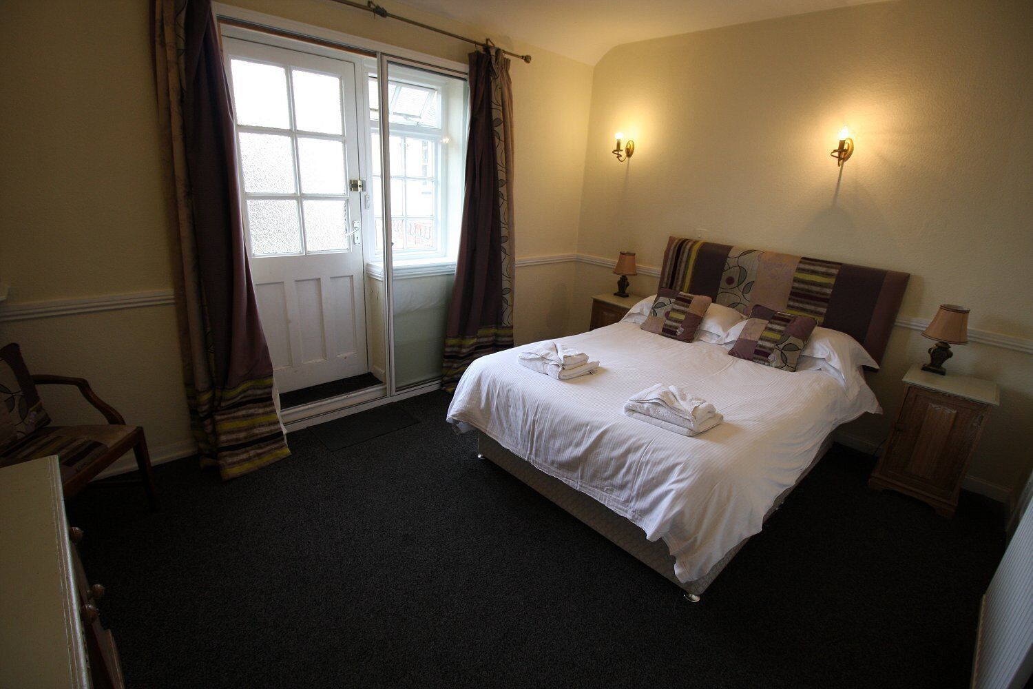 A hotel room with a large bed, two bedside tables with lamps, a door with curtains, and a view of the outside.