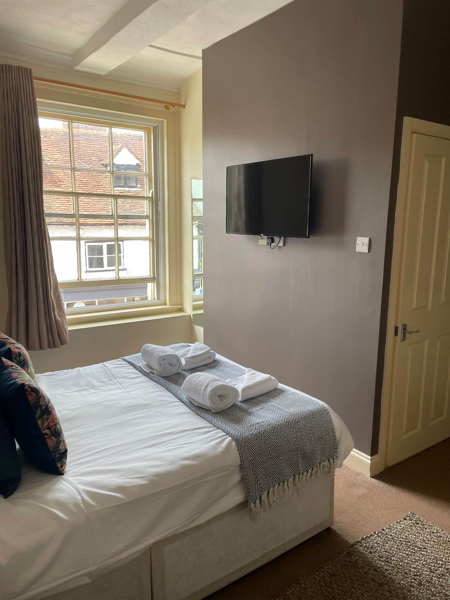 Un dormitorio con una cama, un televisor montado en la pared, una ventana con cortinas y una puerta.