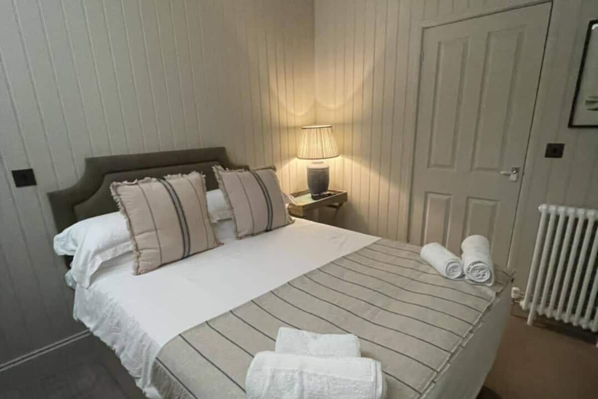 A neatly made bed with a striped blanket and pillows, a bedside lamp, and a hanging light fixture in a room with wooden paneling and a door.