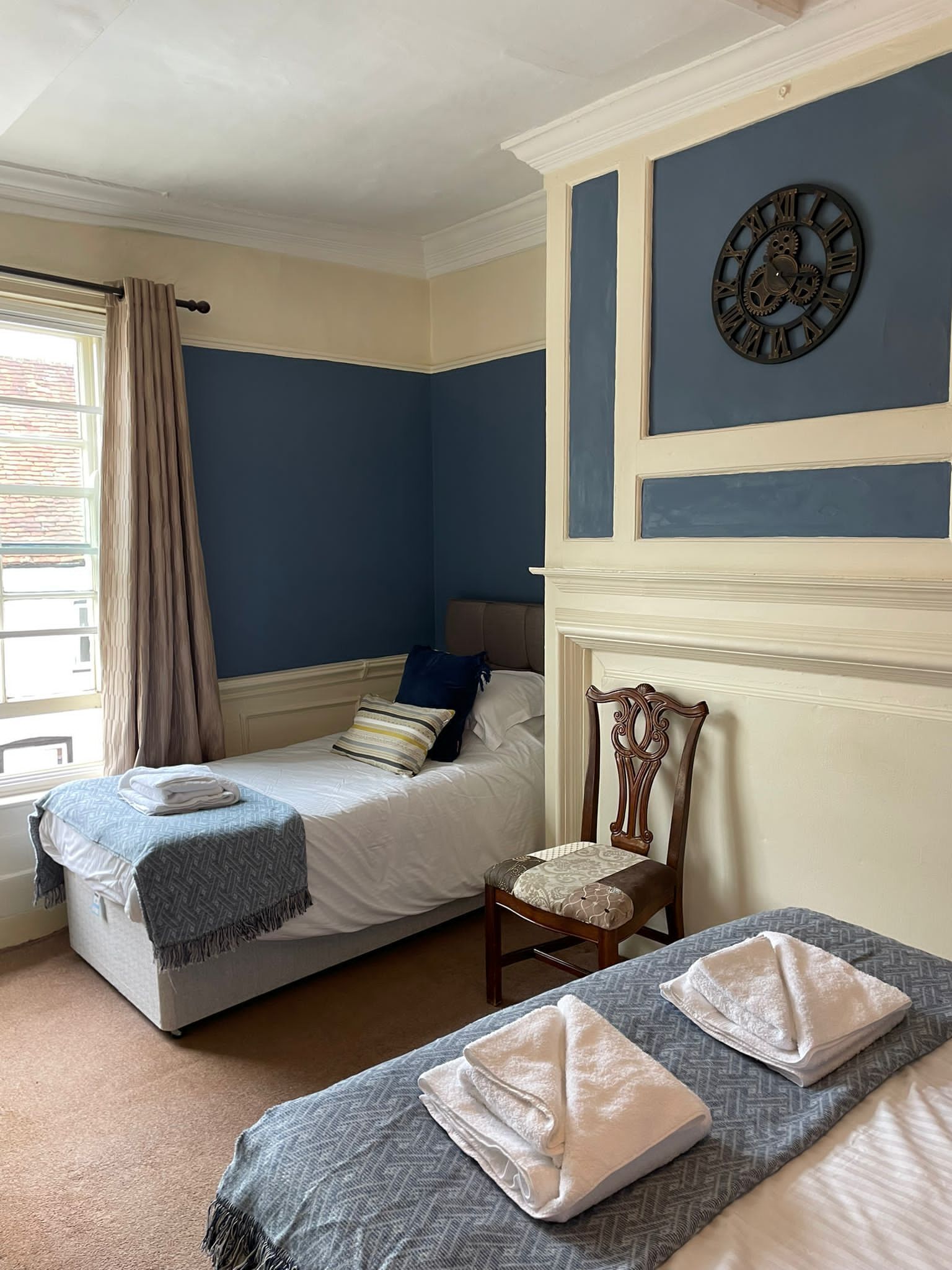 A bedroom with two single beds, a wooden chair, a window with curtains, and a wall clock.