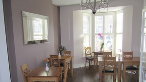 A dining area with a wooden table and chairs, a chandelier, and a window with blinds. - Geminian Guest House (Harrogate)