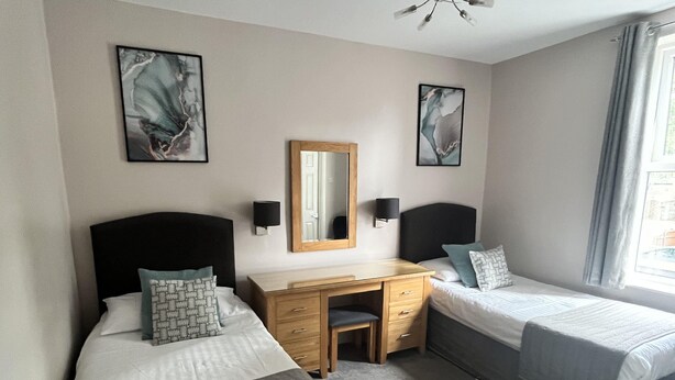 A hotel room with two single beds, a wooden desk, a mirror, and two wall-mounted lamps. - Geminian Guest House (Harrogate)