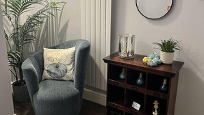 A room with a wooden herringbone floor, a grey armchair with a pumpkin pillow, a small wooden side table with decorative items, a radiator, and a wall-mounted mirror. - Geminian Guest House (Harrogate)