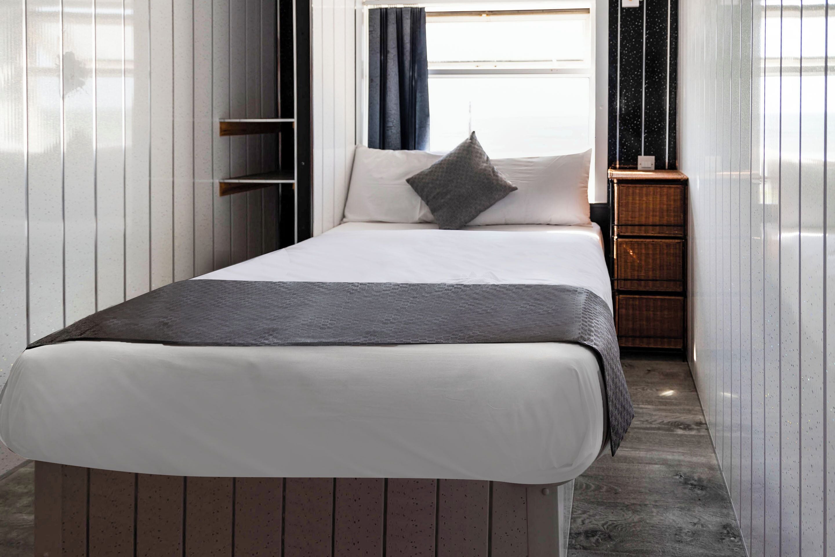 Una cama bien hecha con un colchón blanco y una manta gris, una almohada gris, y una mesita de noche de madera con una cesta tejida a su lado.