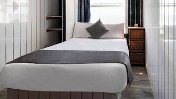Una cama bien hecha con un colchón blanco y una manta gris, una almohada gris, y una mesita de noche de madera con una cesta tejida a su lado.