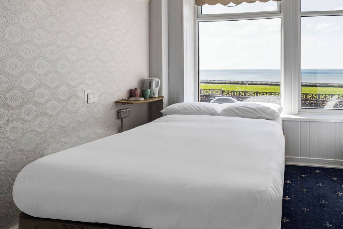 A bedroom with a bed, a window with a view of the sea, a small shelf with a kettle, and a patterned wall.