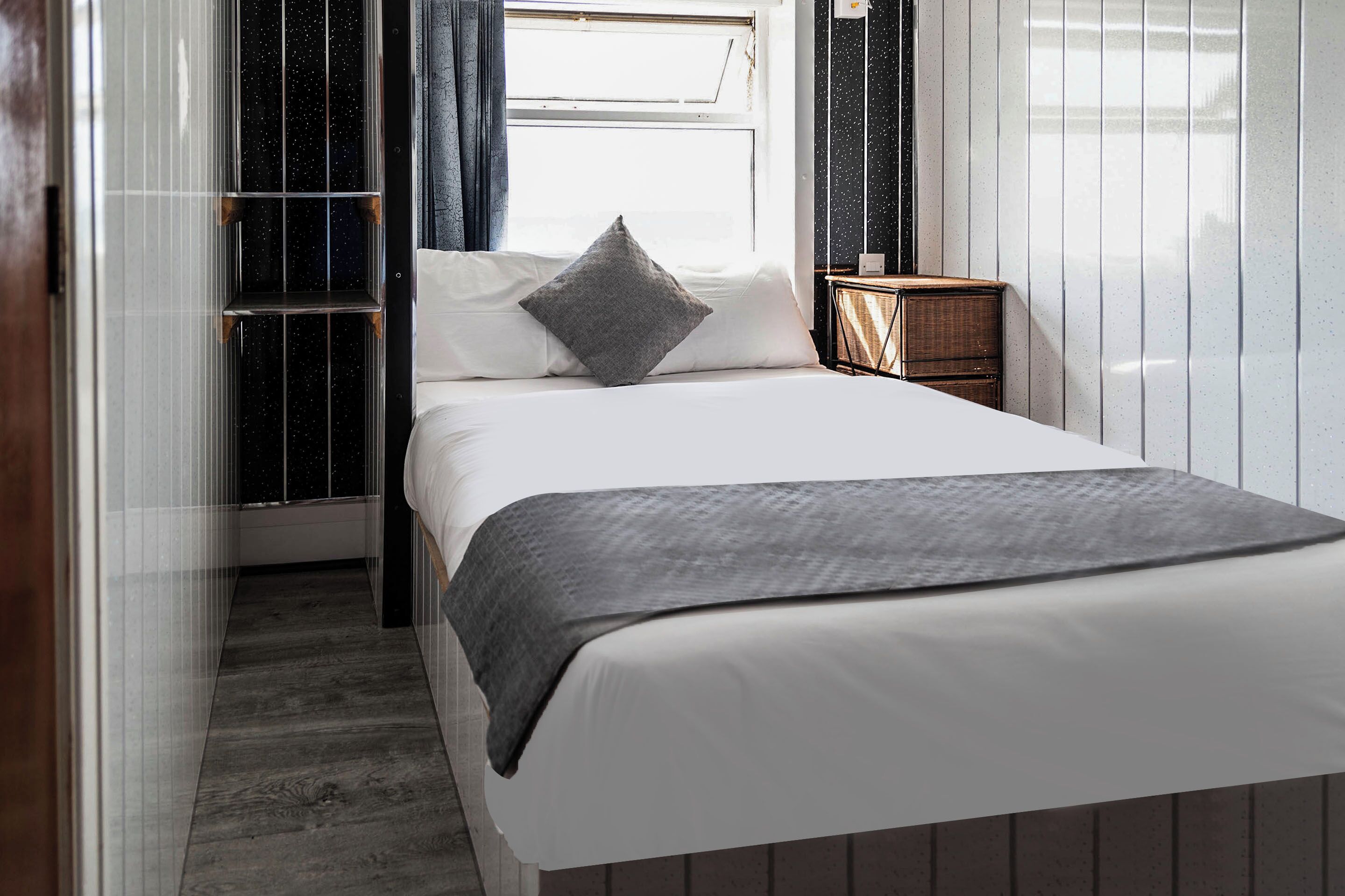 Una cama bien hecha con una manta gris y almohada en una habitación con ventana, una mesita de noche de madera y una pared con líneas verticales.