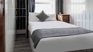 Una cama bien hecha con una manta gris y almohada en una habitación con ventana, una mesita de noche de madera y una pared con líneas verticales.