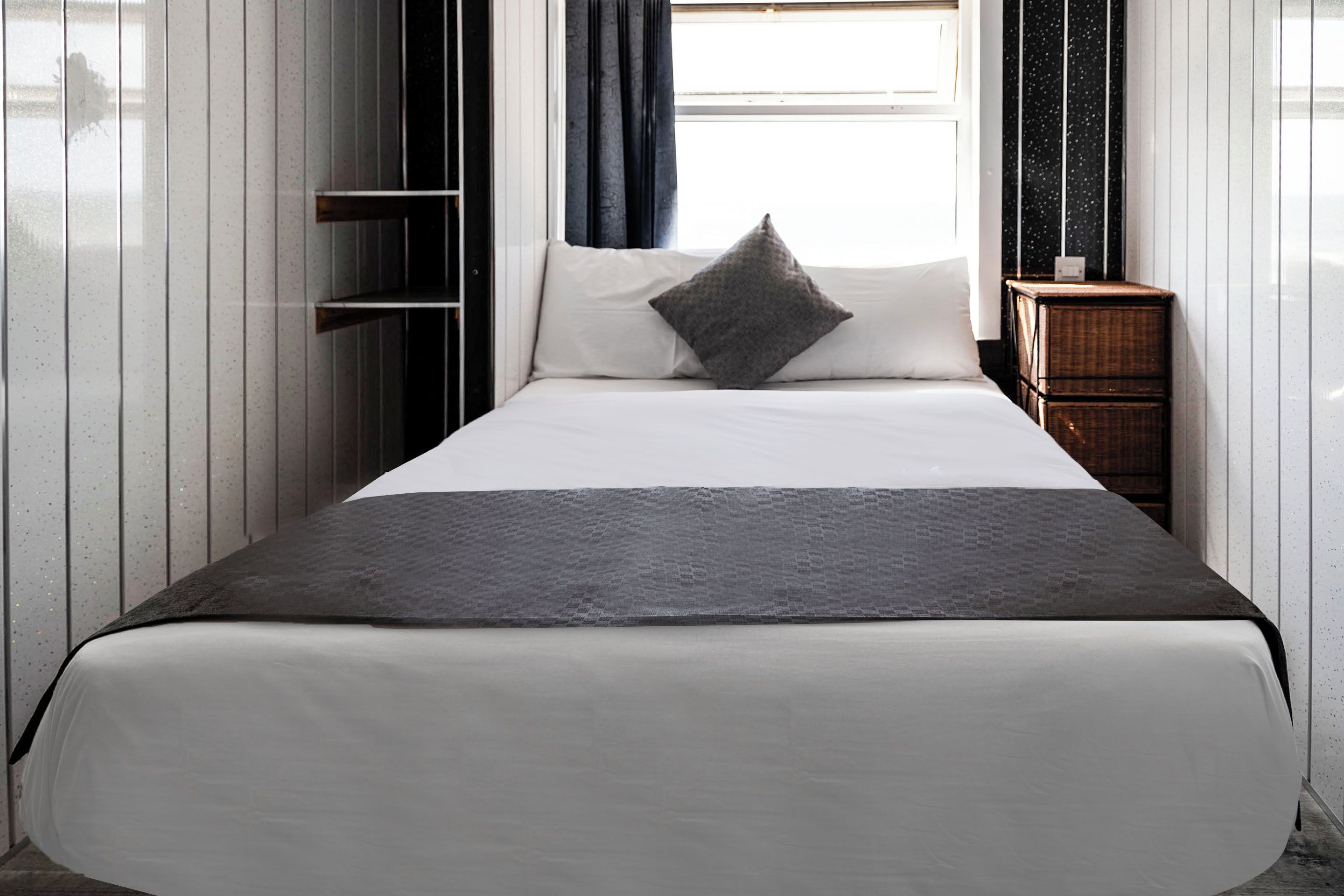 A neatly made bed with a white mattress and a dark gray blanket, a single gray pillow, and a wooden nightstand with a small item on it.