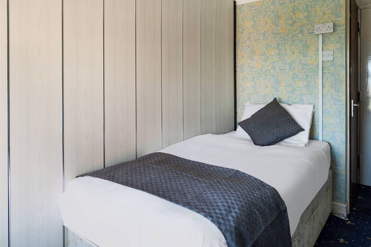 A neatly made bed with a white duvet and a dark grey blanket, positioned against a wall with a light wood panel and a floral wallpaper pattern.
