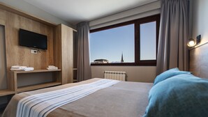 Ein Hotelzimmer mit Bett, Wandfernseher, Fenster mit Blick auf ein Gebäude und Heizkörper.