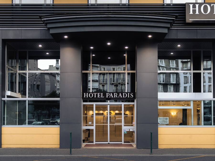 Uma entrada de hotel moderna com fachada de vidro, um letreiro chamativo 'HOTEL PARADIS' e o interior visível pelo vidro.