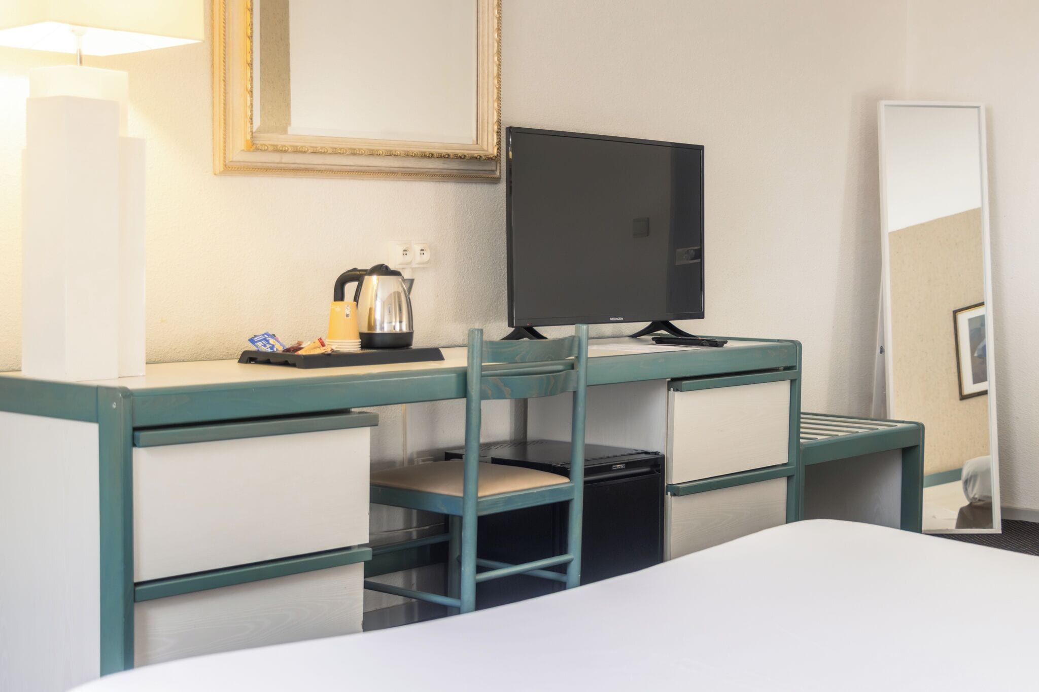 A hotel room with a bed, a desk with a chair, a television, a kettle, and a mirror.