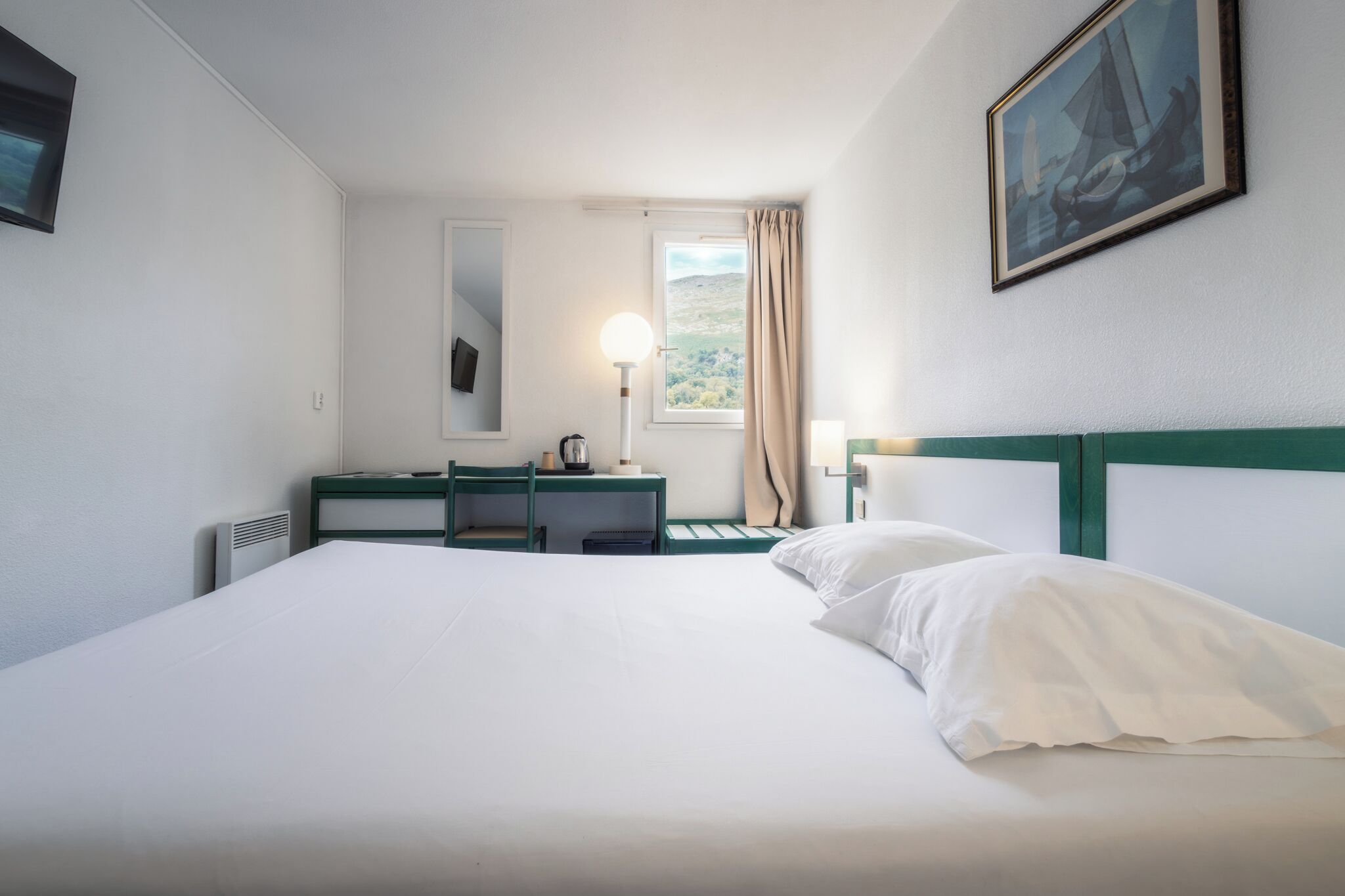 A hotel room with a bed, a desk, a chair, a lamp, a window with curtains, and a painting on the wall.