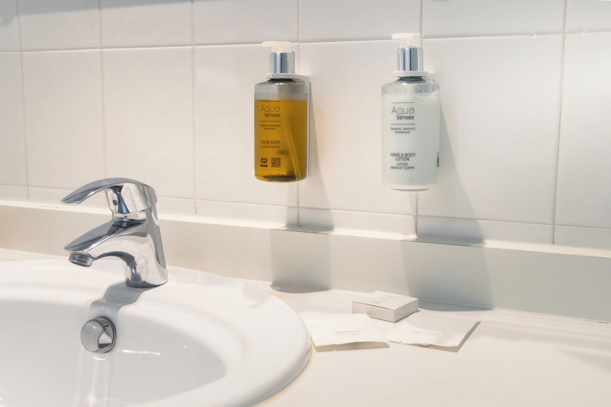 Baño con lavamanos blanco, grifo cromado y dos botellas de jabón líquido Aqua Senses sobre una repisa.