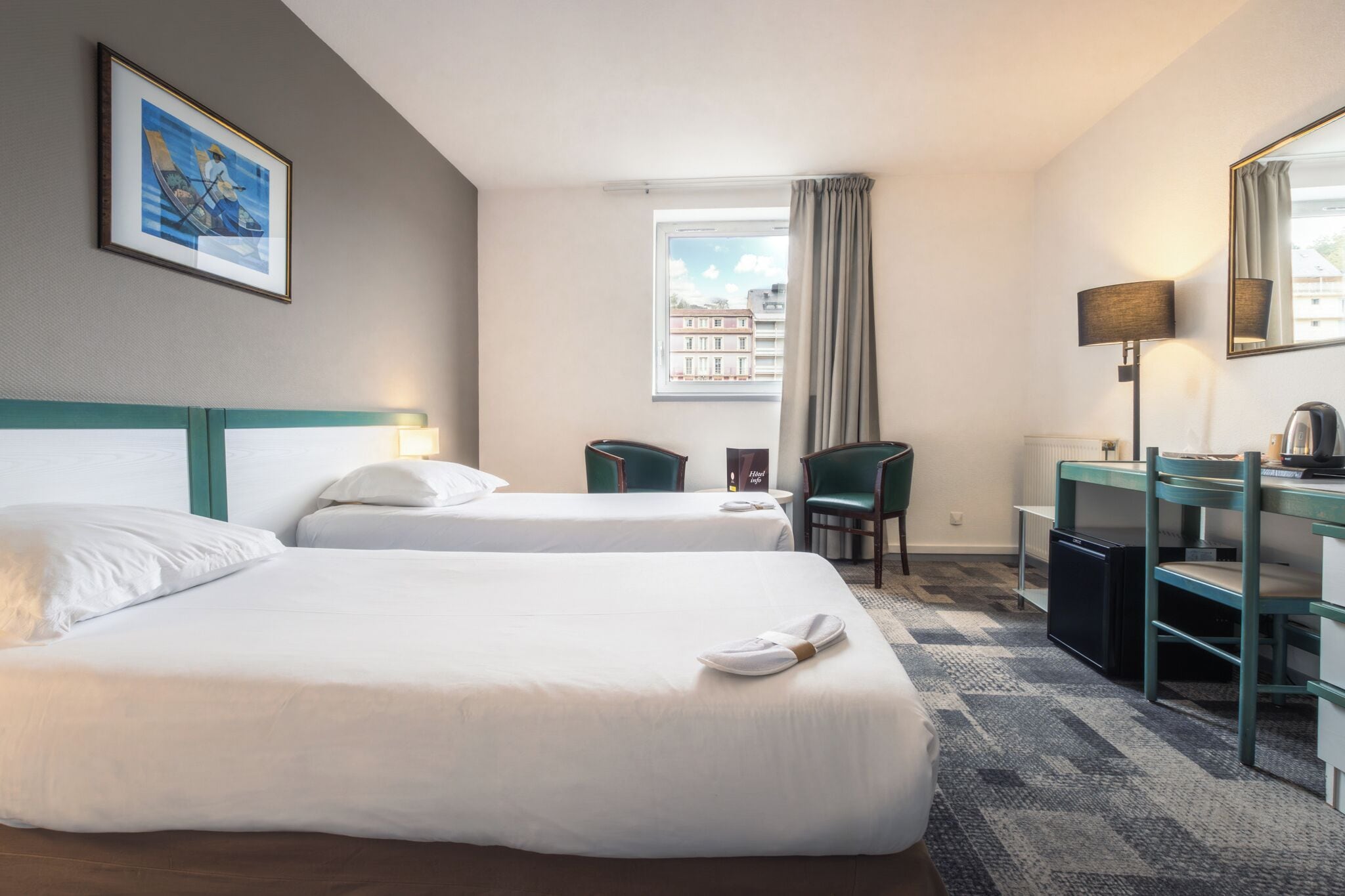 A hotel room with two beds, a desk, a chair, a lamp, a radiator, and a window with a view of buildings.