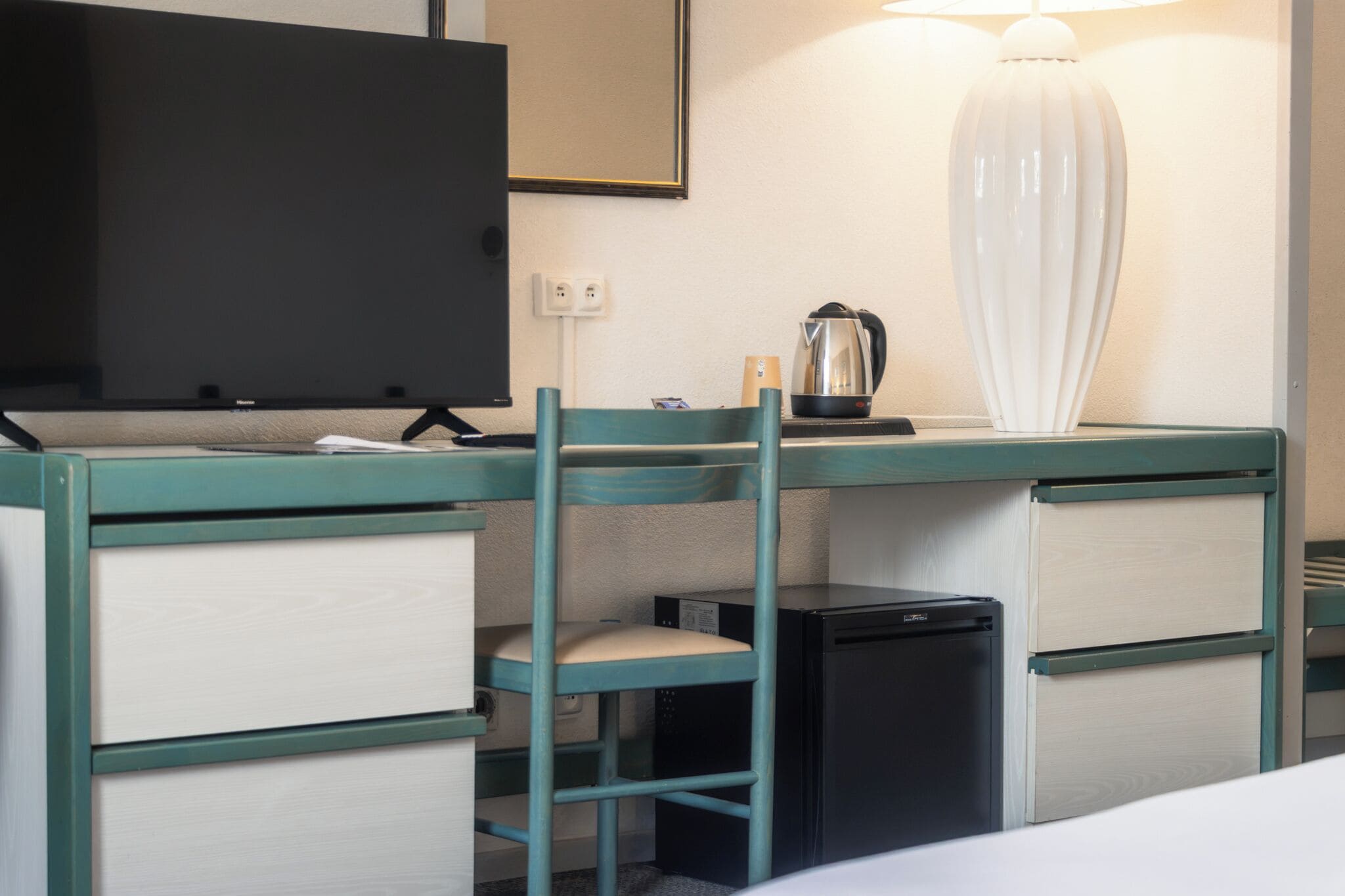 A compact hotel room with a bunk bed, a small refrigerator, a desk with a kettle, a television, and a lamp.