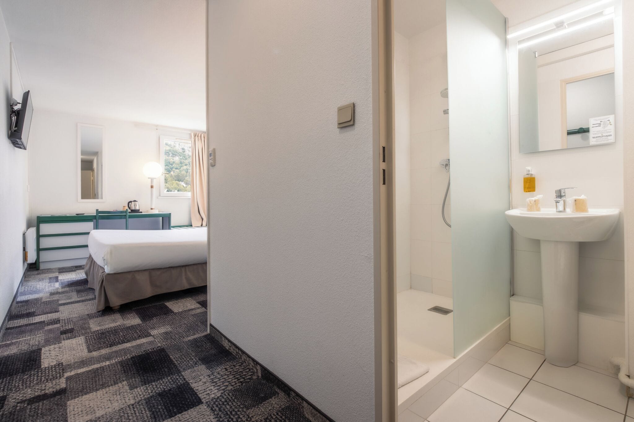 Habitación de hotel con cama, escritorio, espejo, ventana con cortinas y baño con lavamanos y ducha.