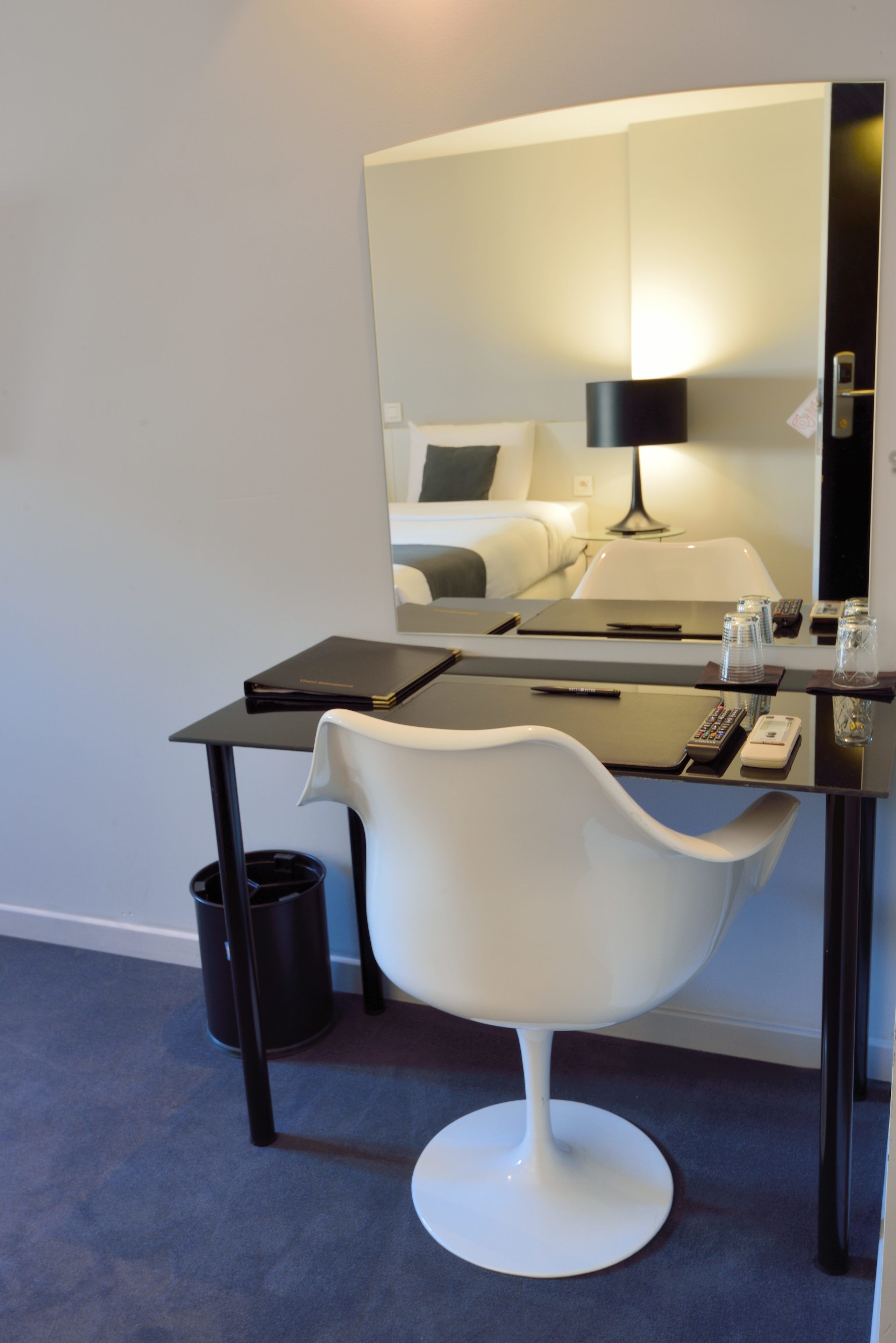 A modern hotel room with a desk, a white chair, a large mirror, and a bed with a white headboard.
