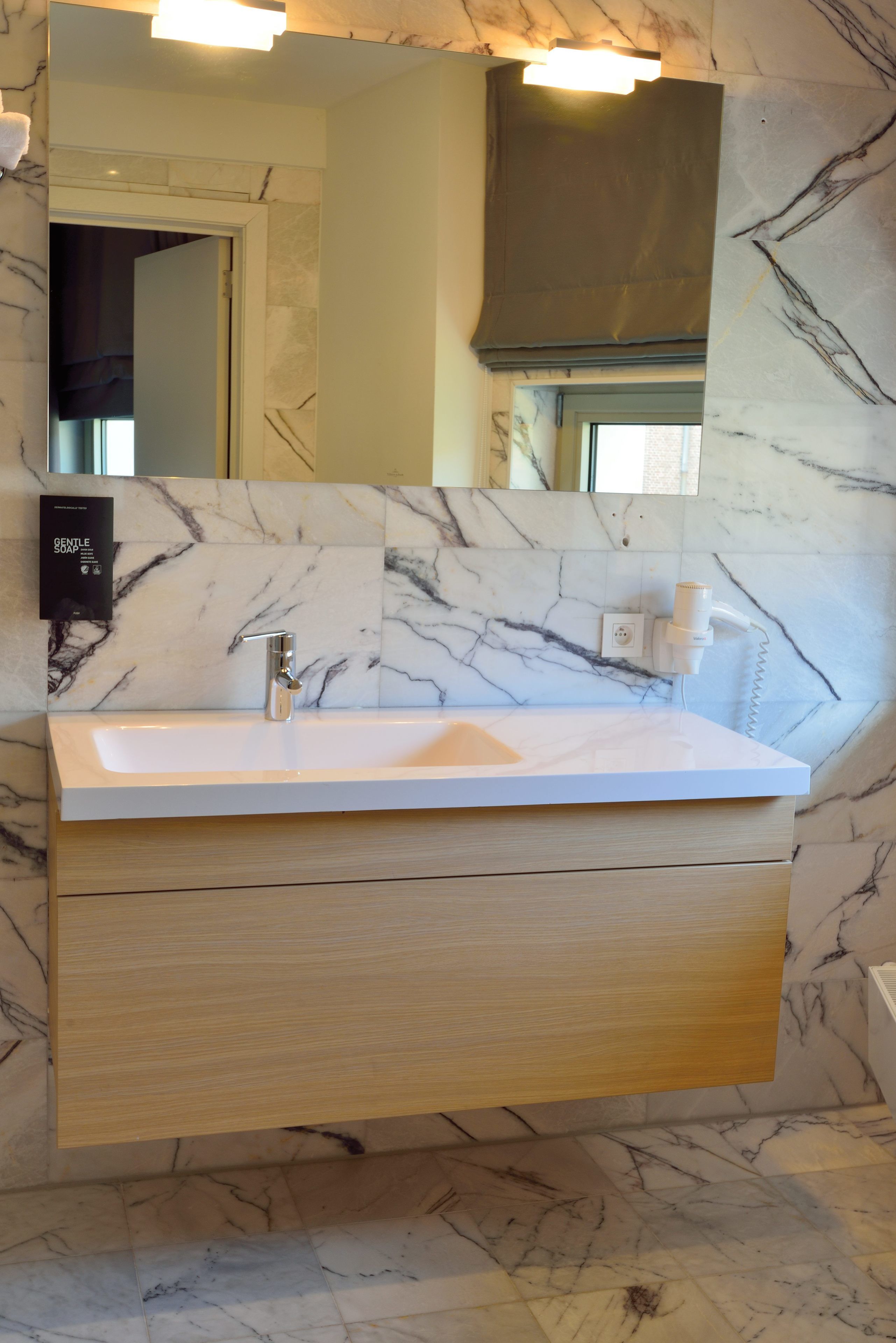 Une salle de bain avec un pare-feu en marbre, un lavabo blanc, un miroir et un meuble en bois en dessous.