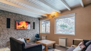 A cozy living room with a stone wall, a fireplace, a TV, and large windows. - Ski Sur Apartments (San Carlos de Bariloche)