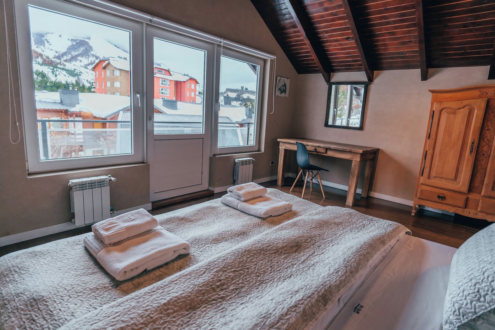 A bedroom with a large bed, a wooden desk, and a view of a snowy mountain and buildings through the windows.