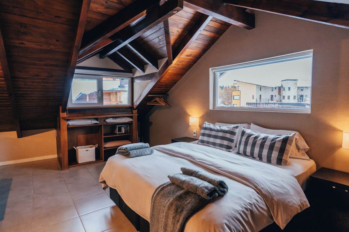 A bedroom with a sloped ceiling, a bed with pillows, a bedside table, a lamp, a window with a view of buildings, and a wooden shelf with a basket.