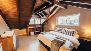 Een gezellig slaapkamer onder het dak, met een schuine plafond, een groot bed met kussens, een nachtkastje, een houten dressoir en een raam met uitzicht op gebouwen.