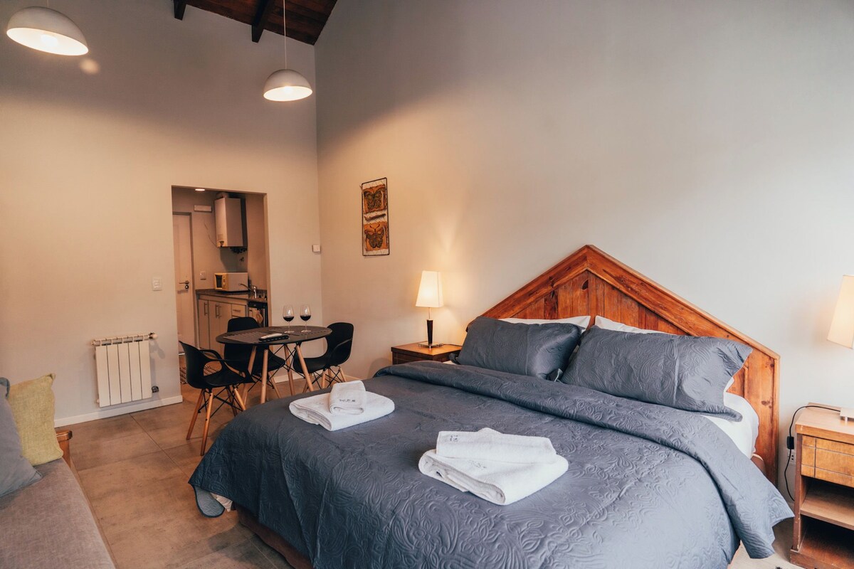 A bedroom with a wooden headboard, a bed with grey bedding, a bedside table with a lamp, and a small table with chairs and a bottle of wine.