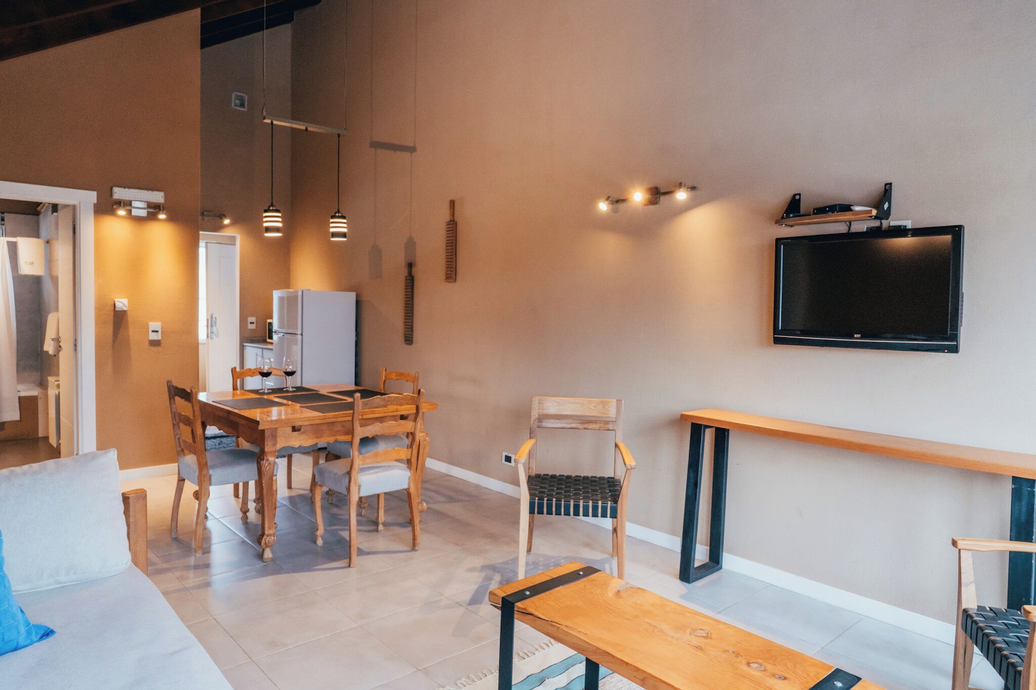 A modern living room with a dining area, a flat-screen TV mounted on the wall, wooden furniture, and pendant lighting.