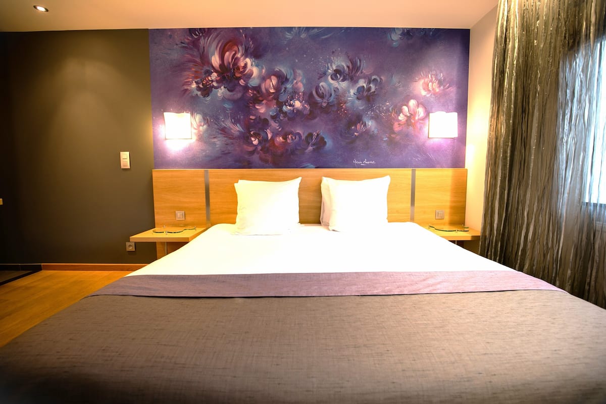 A hotel room with a large bed, two bedside tables, a headboard with a painting, and a curtain on the window.