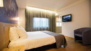 Ein Hotelzimmer mit Bett, Fernseher an der Wand, Stuhl und Fenster mit Vorhängen.